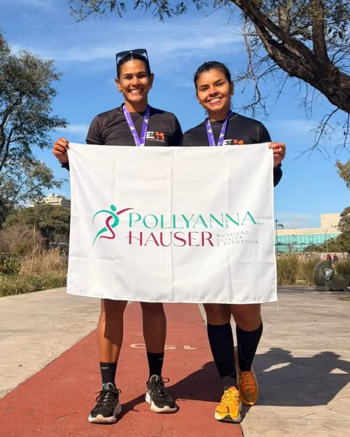 Duas atletas acompanhadas por Pollyanna Hauser segurando a bandeira da marca, com medalhas no pescoço.